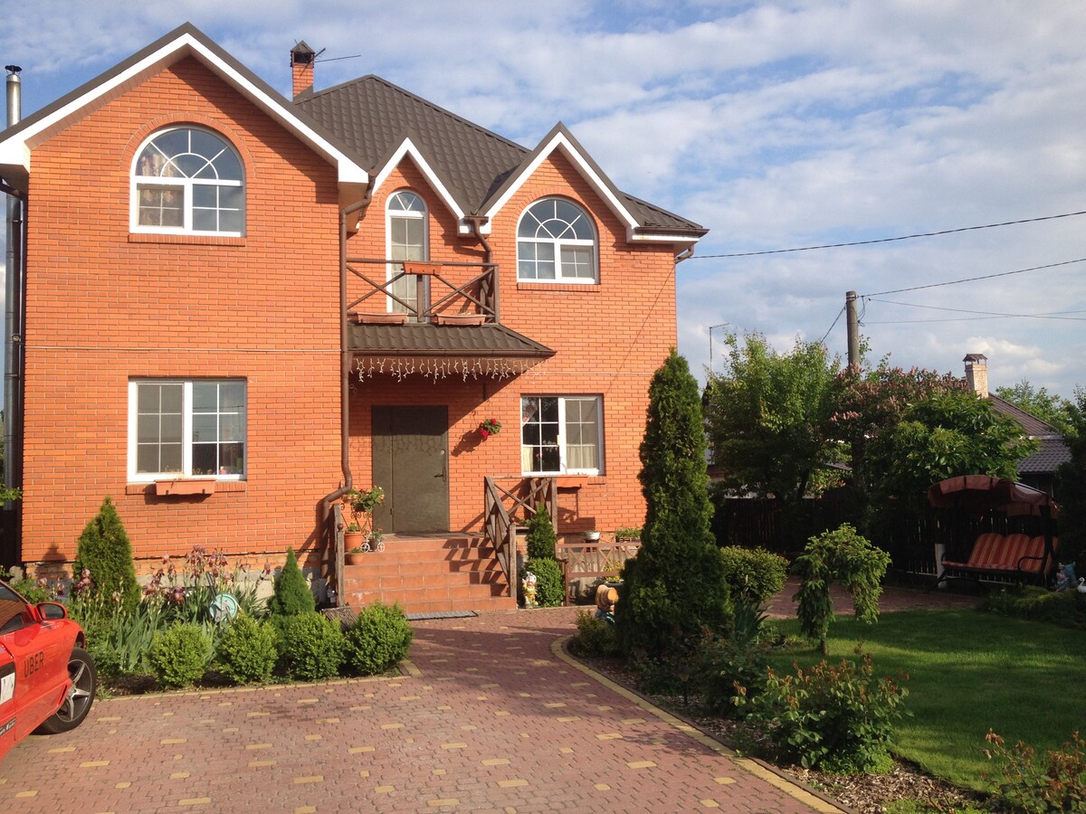 A two-story brick house features large arched windows and a welcoming entrance. The well-maintained garden includes neatly trimmed shrubs and flowers. A patio area with seating is visible, providing a relaxing outdoor space for guests.
