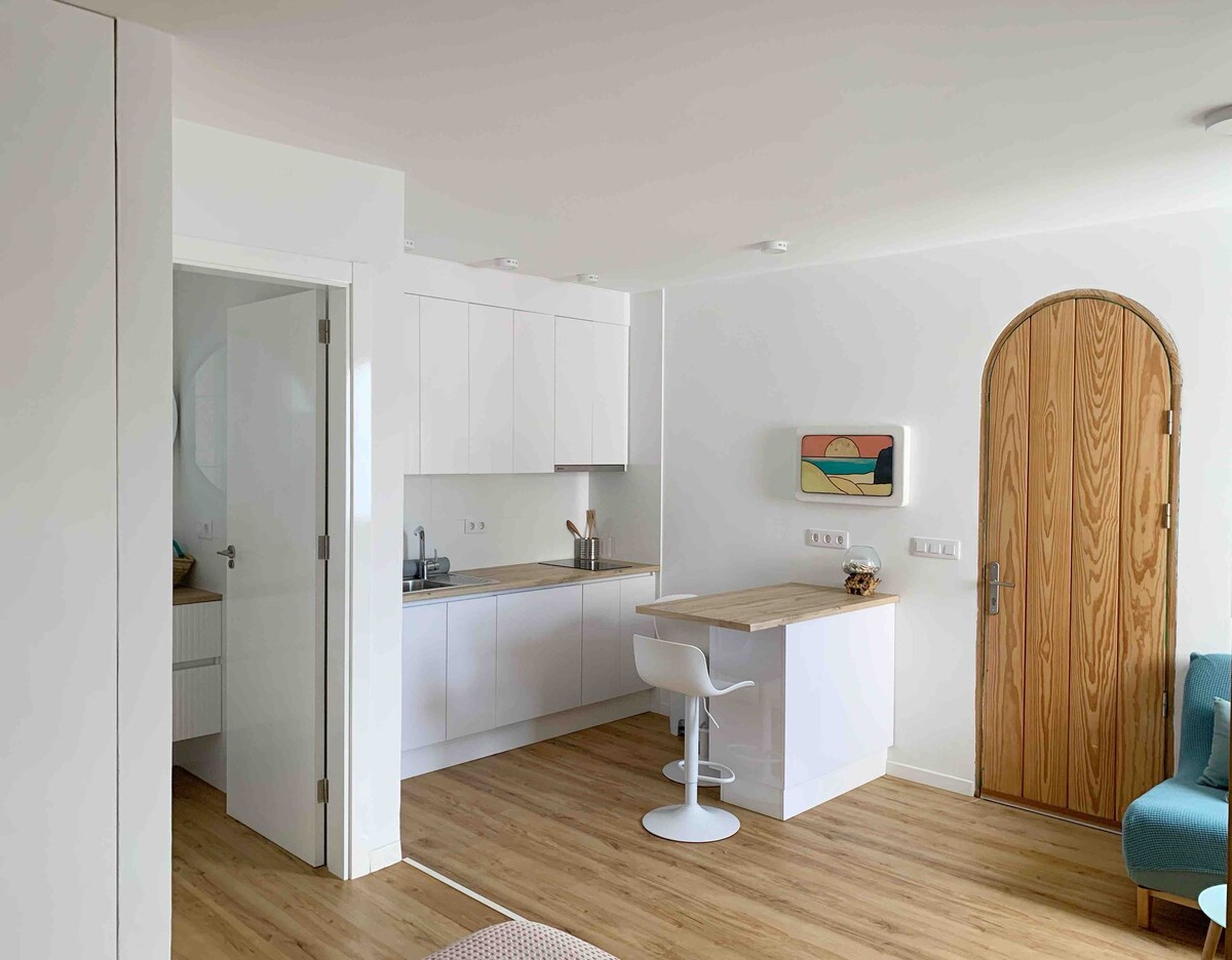 A modern kitchen area is paired with a small bar featuring a streamlined counter and a contemporary white stool. The space is brightened by natural light from the nearby entrance, which includes a wooden door with an arched design.