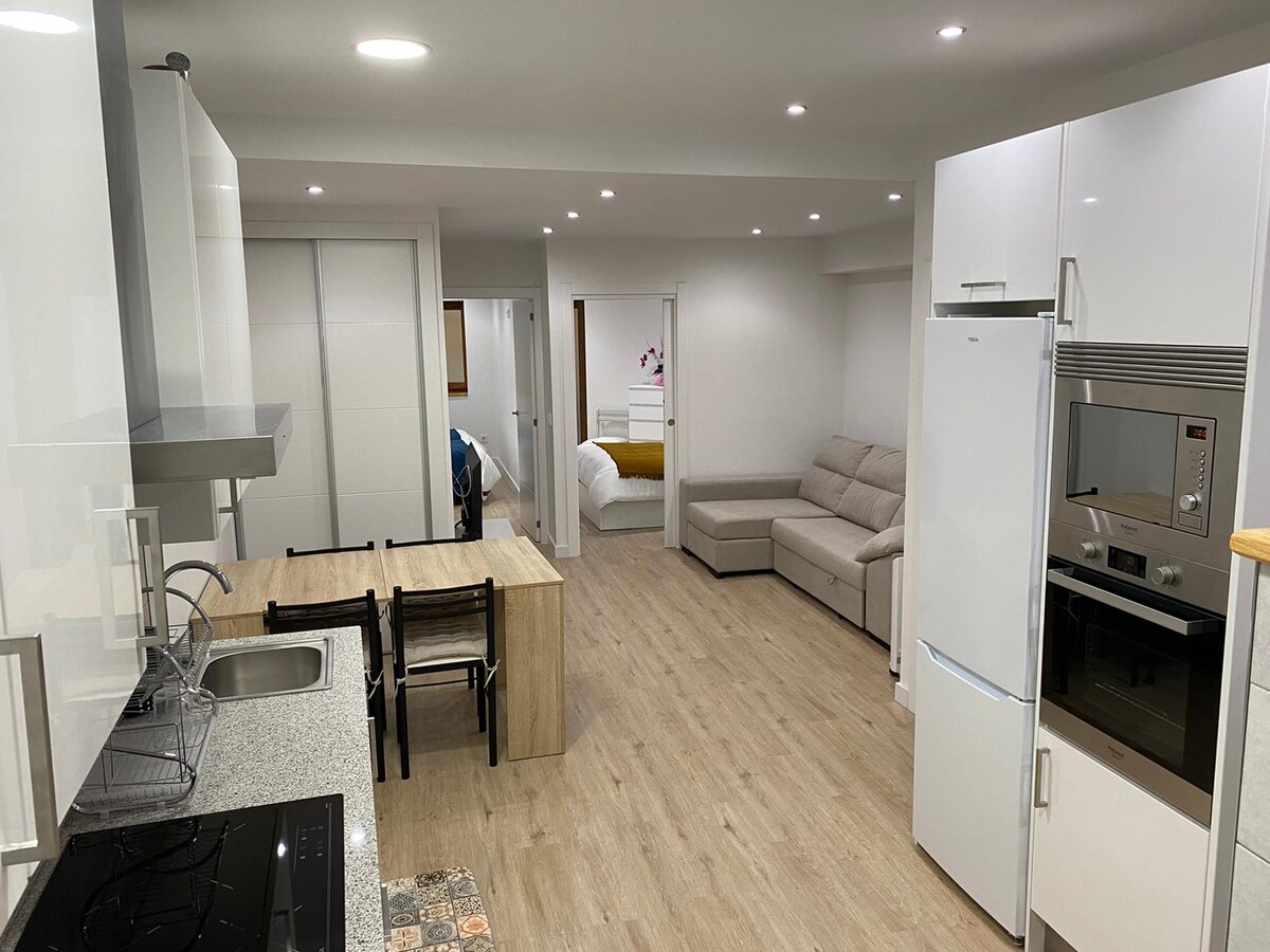 A modern kitchen area features sleek cabinetry and a spacious countertop. A dining table with four chairs is positioned nearby, and a comfortable sofa is visible in the living space. The open layout allows natural light to brighten the space, enhancing the welcoming atmosphere.