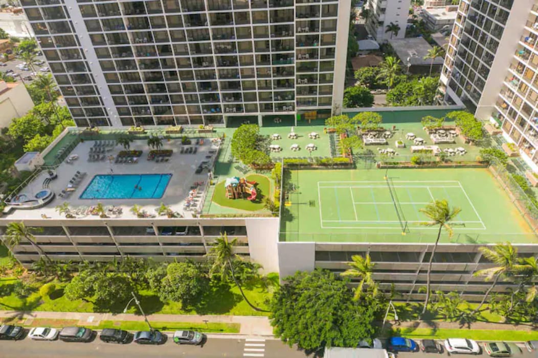 An aerial view showcases the 6th-floor recreation deck of Waikiki Banyan. A large heated pool is surrounded by lounge chairs, with a children's play area visible nearby. A tennis court is located adjacent to the pool, blending seamlessly with lush greenery and palm trees.