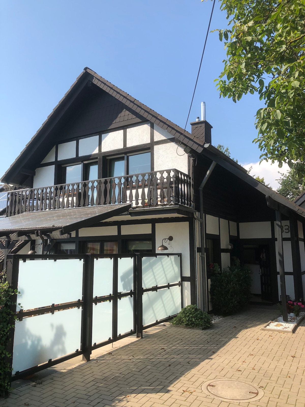 The exterior of a charming two-story house is presented, featuring a dark wood and white panel design. A covered balcony with a decorative railing is located above the entrance. Lush greenery and a paved pathway lead to the entryway, highlighting the inviting facade.