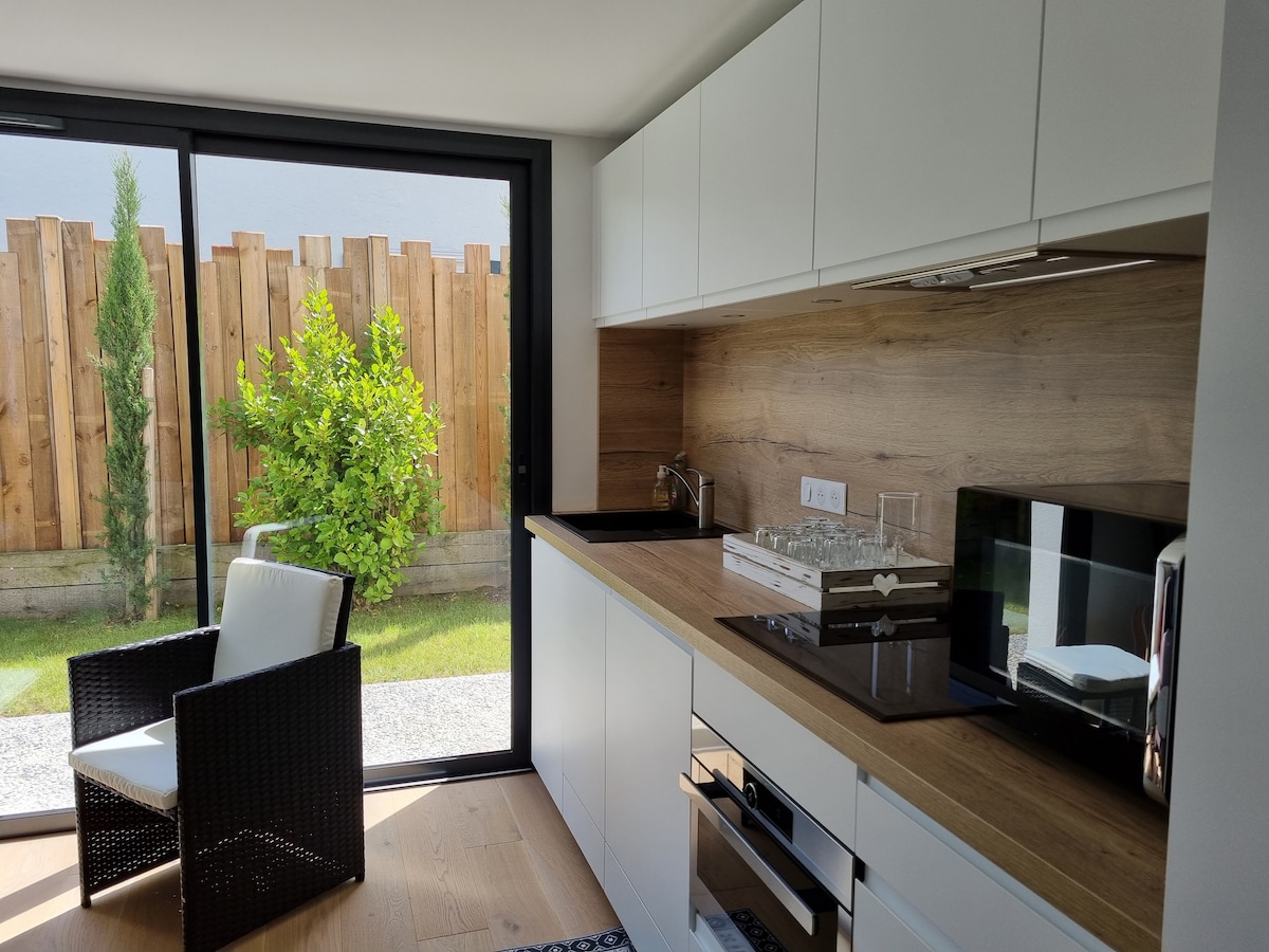 A modern kitchen area features sleek white cabinets against a warm wood backdrop. A microwave and a sleek stovetop are visible. Large glass doors provide a view of a private garden with greenery, enhancing the space's natural light.