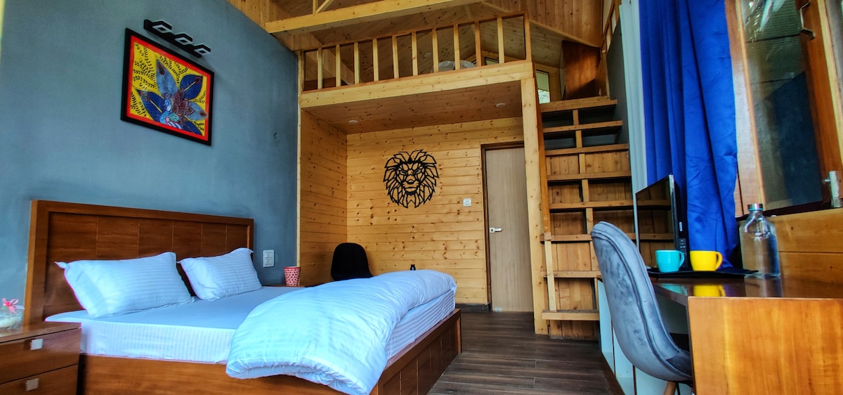 A wooden interior features a spacious bed with white linens and a cozy seating area. A lofted sleeping space is visible above, accessed by a wooden staircase. Natural light floods the room through blue curtains, complemented by artistic wall decor and a desk with colorful mugs.