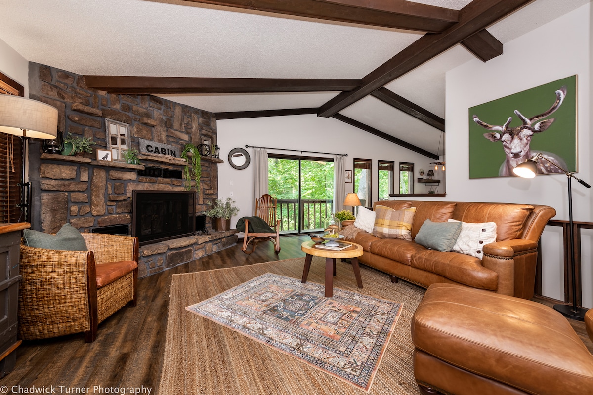 A cozy living area features a stone fireplace, accented by a wooden mantel and decorative elements. Comfortable seating includes a leather sofa and a woven chair. Natural light is welcomed through large windows, offering views of the surrounding greenery, while a patterned area rug adds warmth.