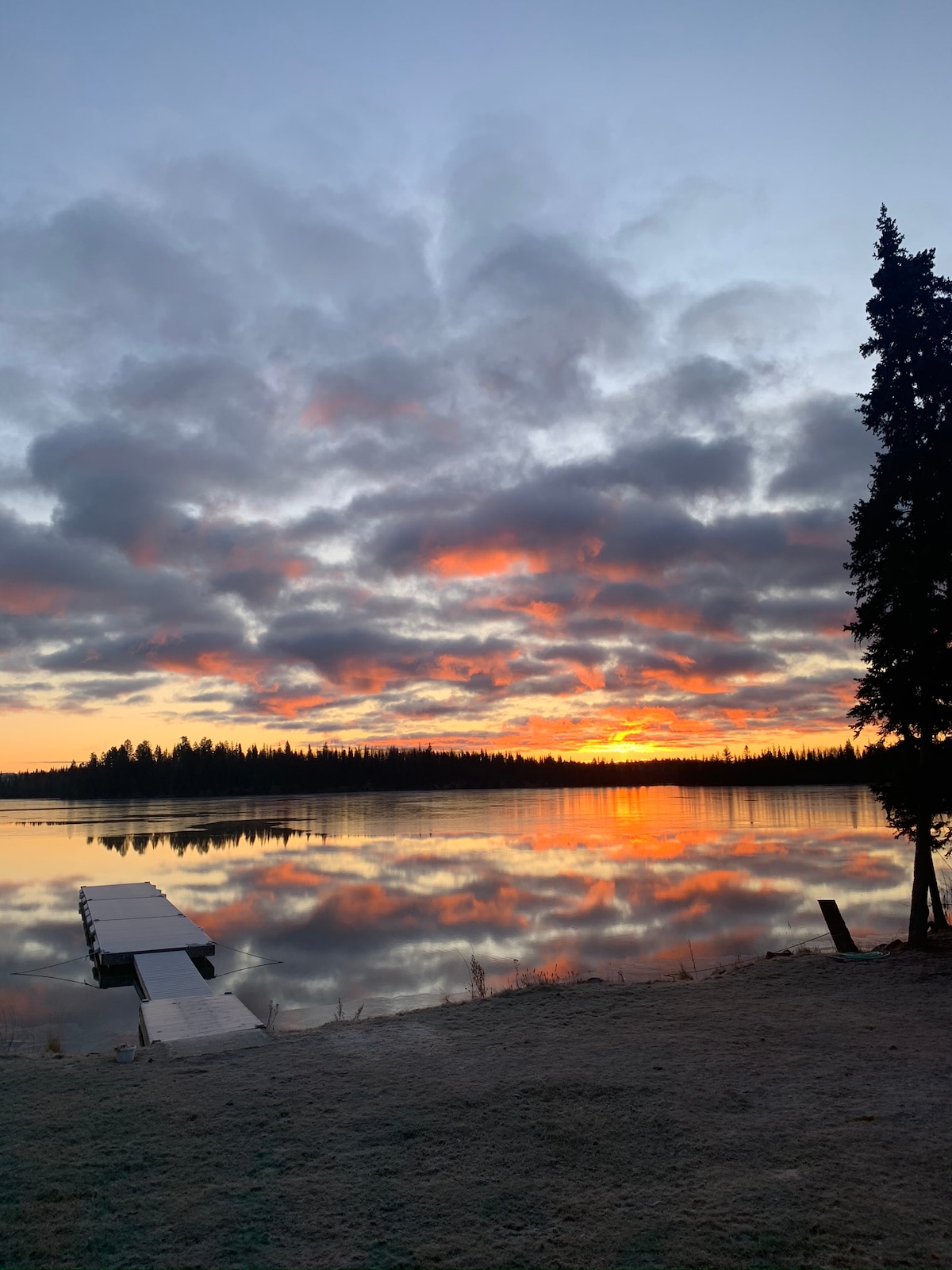 A serene lakeside view showcases a vibrant sunset reflecting off the calm waters. Fluffy clouds in shades of orange and pink contrast against the darkening sky. A wooden dock extends into the lake, surrounded by tranquil natural scenery.