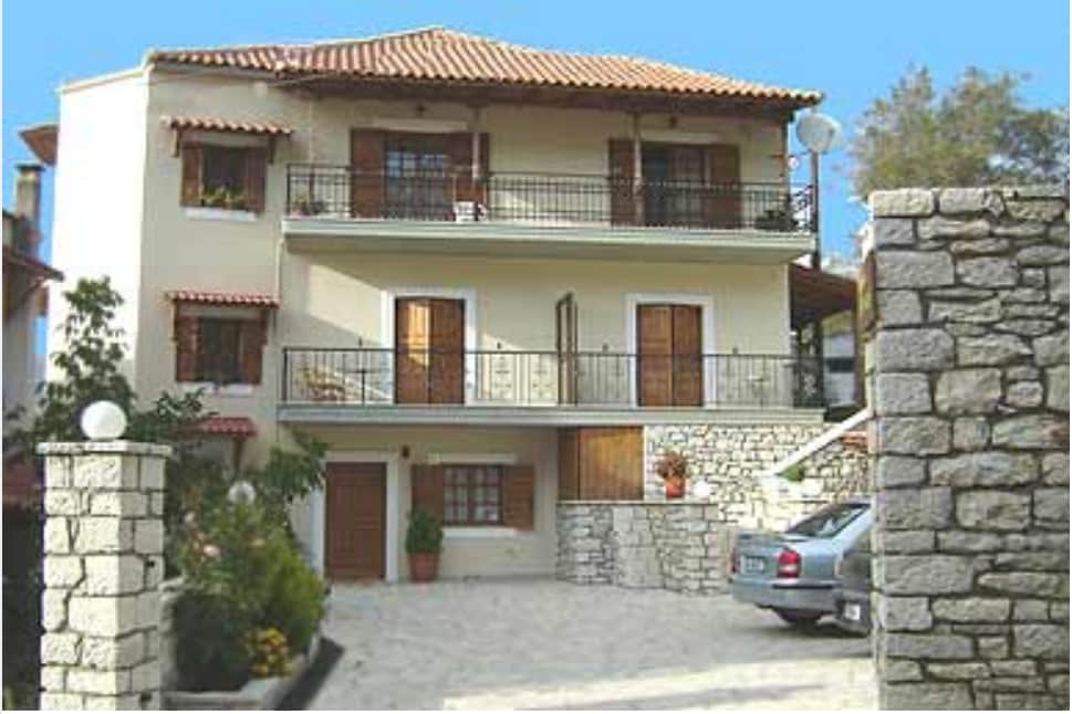 The exterior of the property is shown, featuring a three-level structure with multiple balconies and wooden shutters. A stone wall encloses the entrance, leading to a paved driveway with parking space. Lush greenery and potted plants add warmth to the welcoming facade.