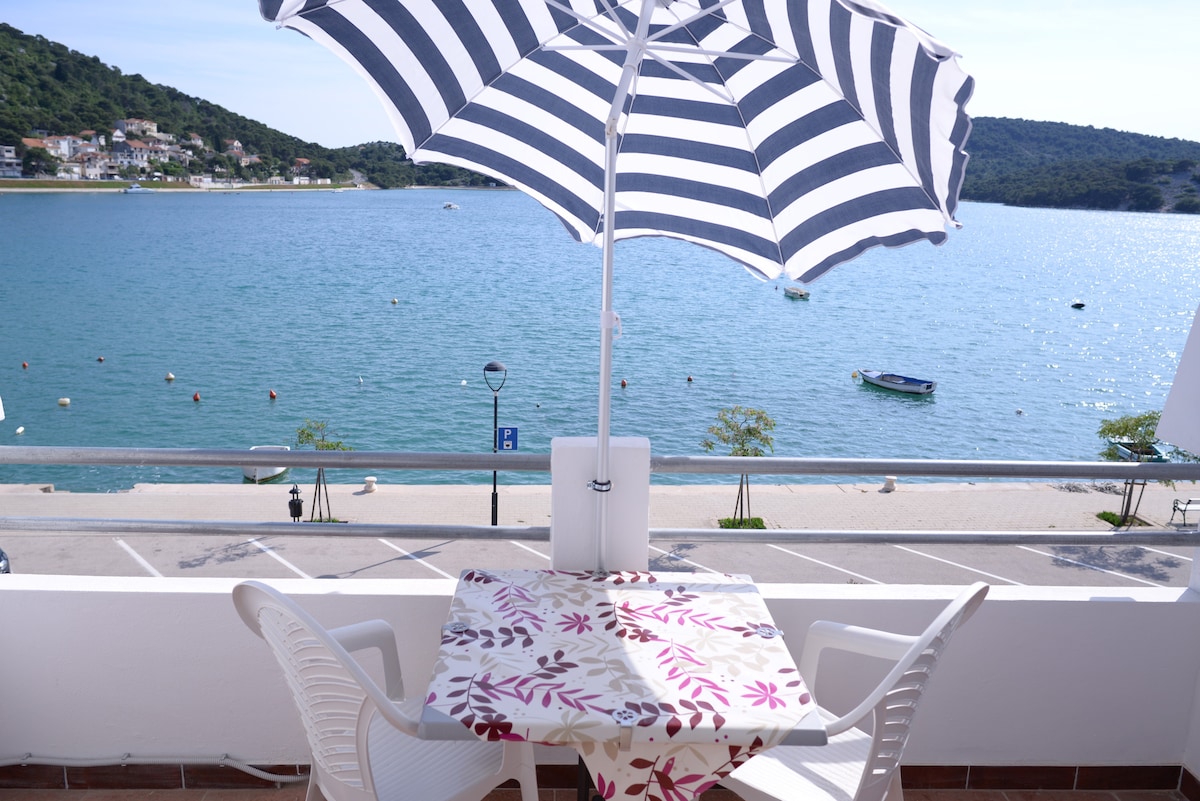 A spacious seaview balcony features a table covered with a patterned cloth and two white chairs. A large striped umbrella provides shade, while the tranquil water and distant shoreline create a scenic backdrop for outdoor relaxation.