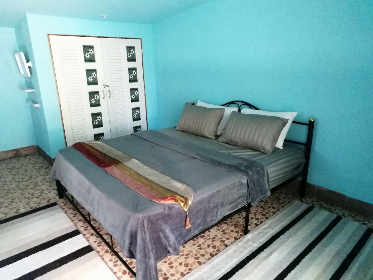 A cozy sleeping area is featured, with a spacious bed dressed in gray linens and accent pillows. The walls are painted a calming shade of turquoise. A patterned rug complements the floor, and storage is provided by the built-in wardrobe visible in the background.