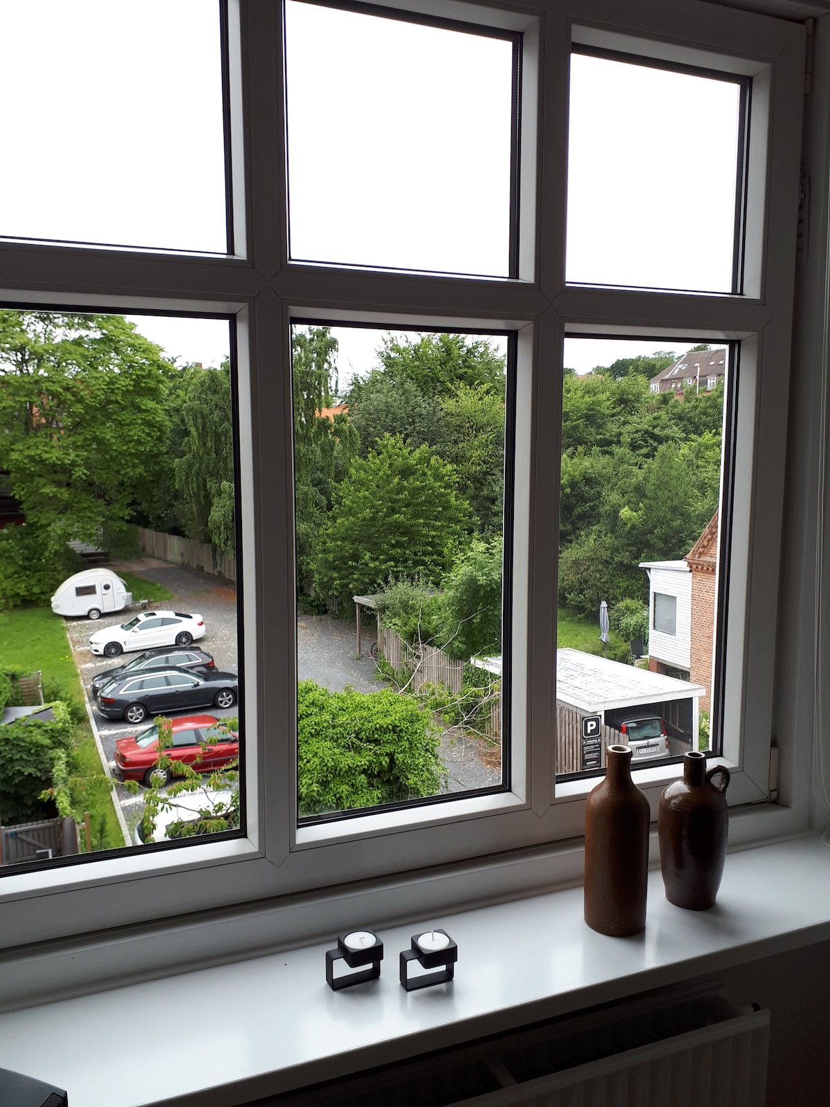 A large window provides a view of the surrounding green area and a parking lot. Two candles are placed on the windowsill, alongside two ceramic vases. The scene highlights the peacefulness of the neighborhood and the greenery outside.