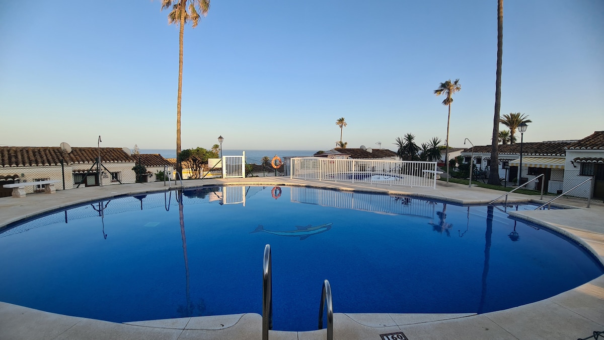A serene pool area features a smooth, circular design with clear blue water reflecting the sky. Surrounding palm trees provide shade, while the distant ocean can be seen just beyond the poolside. A safety fence runs along the perimeter, enhancing the inviting space.