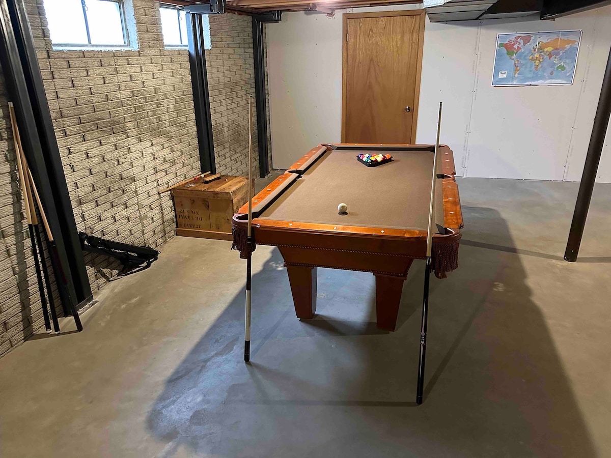 A well-maintained basement features a spacious pool table, with cues placed against the wall and a wooden box nearby for storage. Natural light enters through several windows, illuminating the neutral grey flooring and brick wall, creating an inviting space for entertainment.