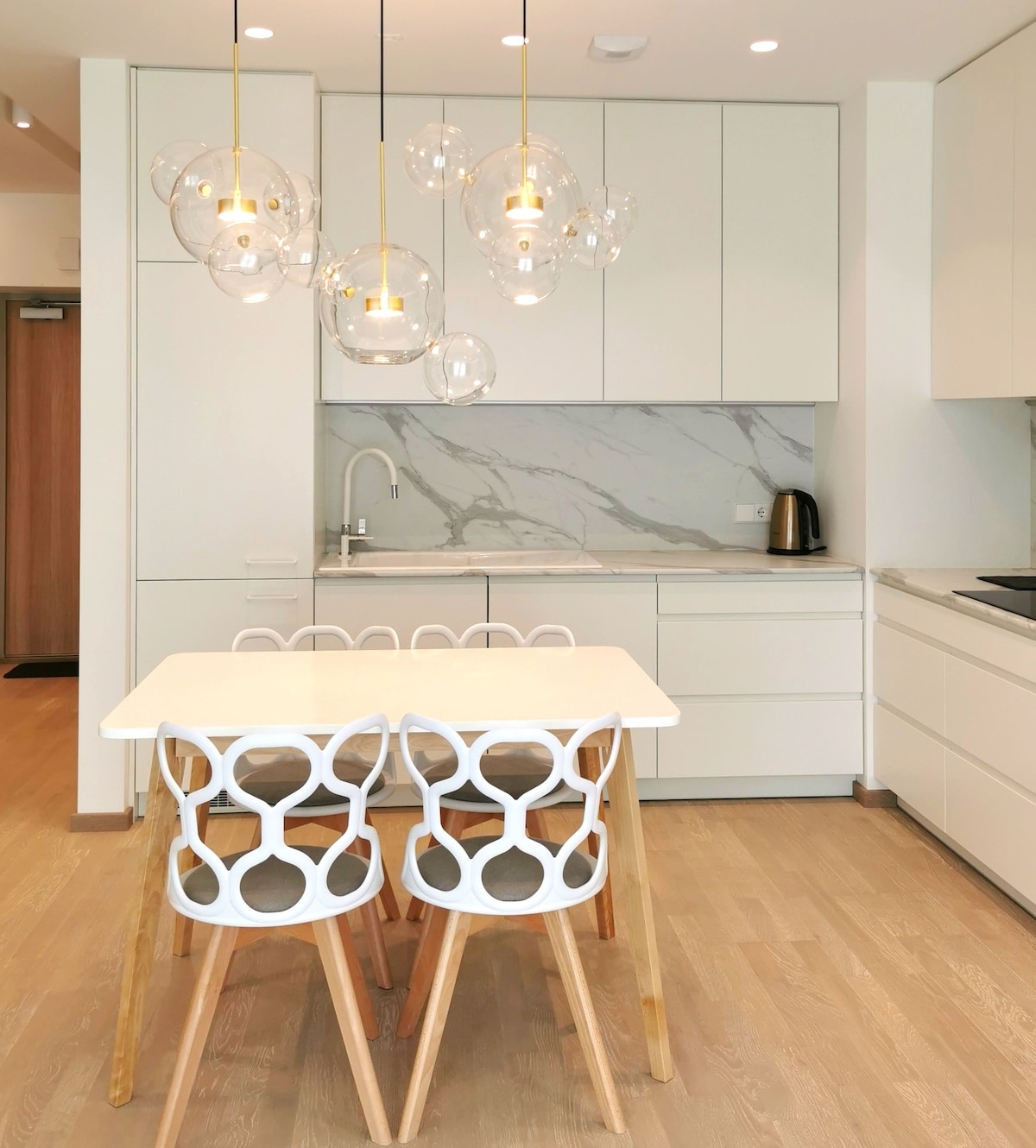 A modern kitchen is featured, showcasing sleek white cabinetry and a marbled backsplash. A round dining table is surrounded by four distinctive chairs with circular cutouts. Unique pendant lights provide illumination above the table, enhancing the clean and stylish design.