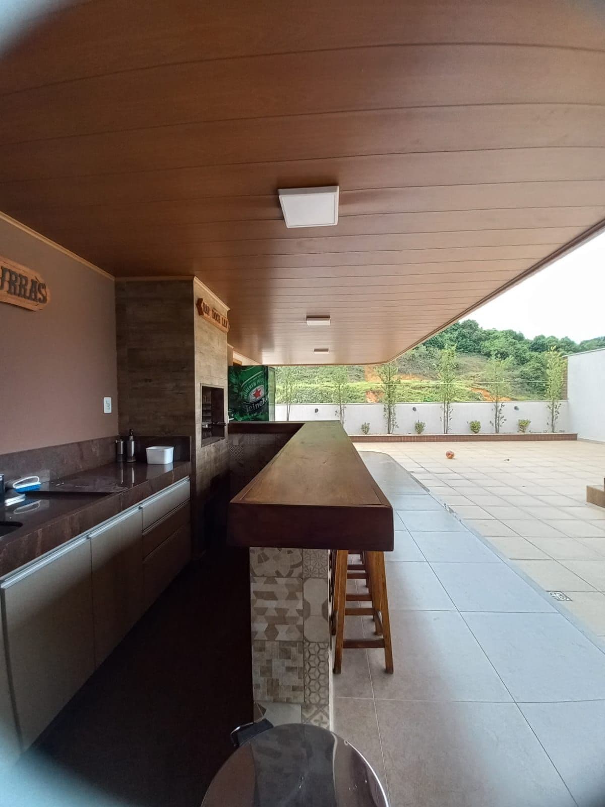 An outdoor area features a sleek bar with a wooden countertop and a stone back wall, set against a backdrop of greenery. The space is well-lit by recessed ceiling lights, and large tiles cover the ground, contributing to a clean and open atmosphere.