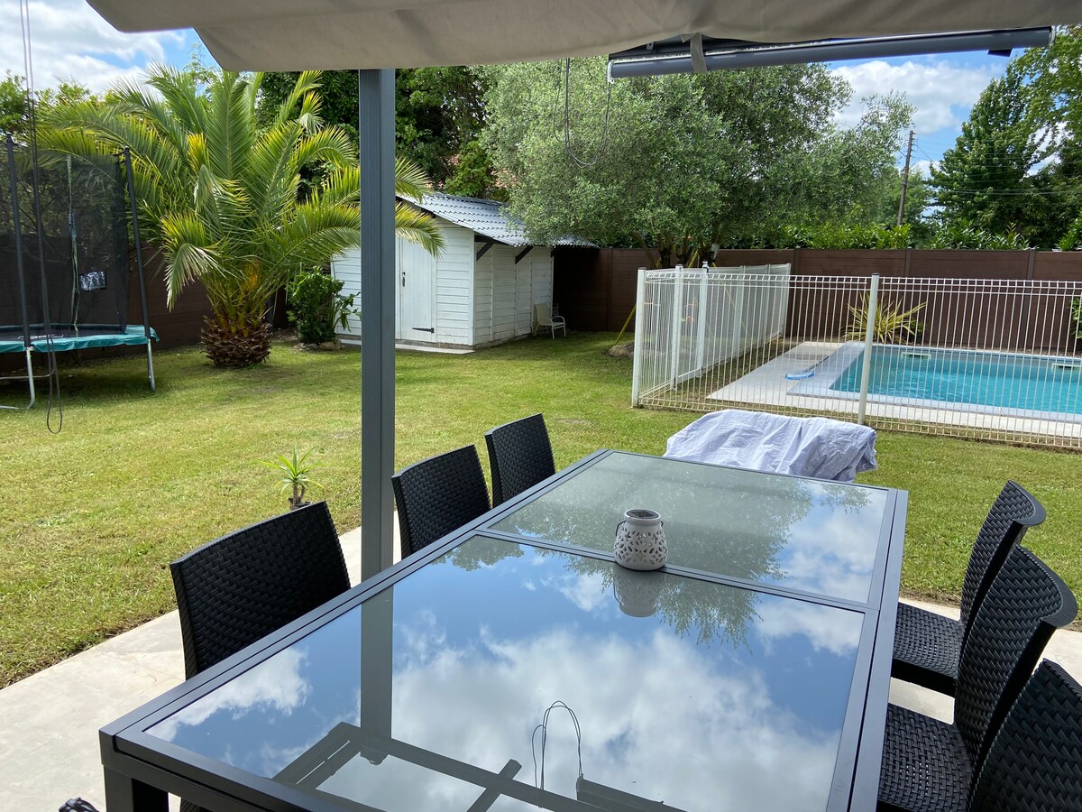 An outdoor dining area features a glass table surrounded by black wicker chairs, set under a shaded canopy. A salt-water pool is visible in the background, along with a fenced garden hosting a trampoline and a storage shed, framed by trees and greenery.