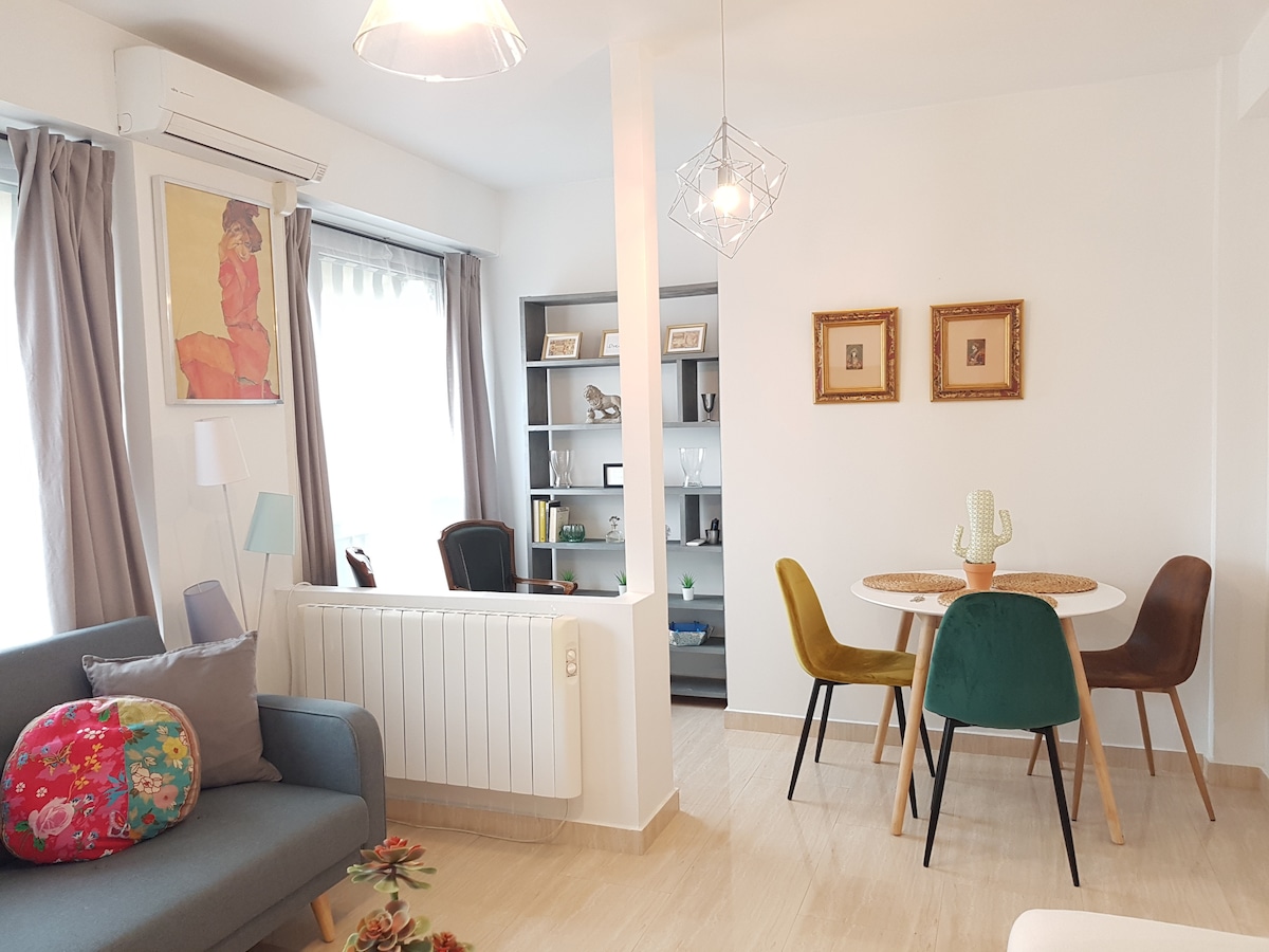 A bright living space is featured with a comfortable sofa adorned with a colorful cushion. A dining area with a round table and stylish chairs is visible, complemented by a wall shelf displaying decorative items. Natural light filters through the large windows, enhancing the inviting atmosphere.