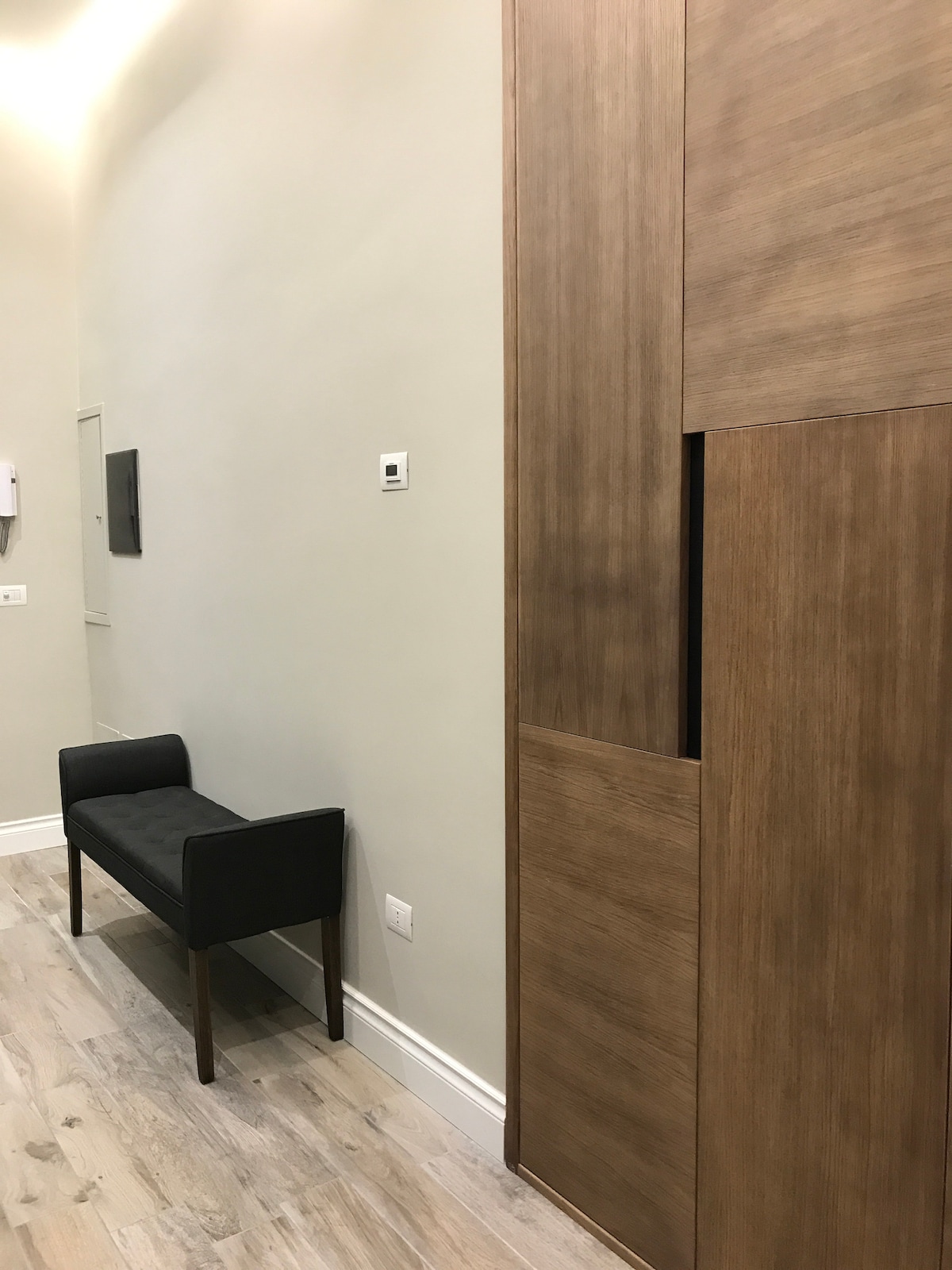A sleek hallway features a light-colored wall and warm wooden paneling. A simple black bench is positioned against the wall, accompanied by a subtle storage unit. Soft lighting enhances the modern design, creating a welcoming entry space.