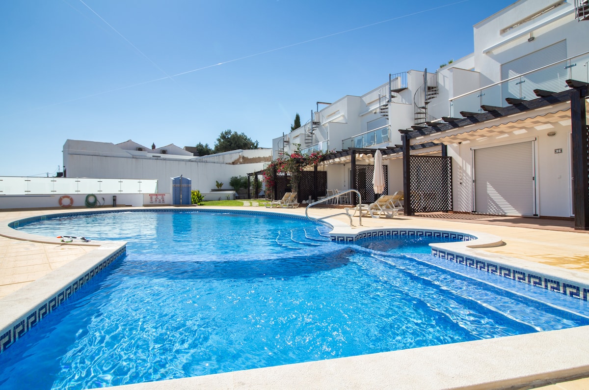 A shared pool is surrounded by a sunlit pool deck, featuring lounge chairs arranged neatly along the edge. The smooth water reflects the clear blue sky, while the surrounding buildings offer a glimpse of a well-maintained community with greenery and vibrant flowers.