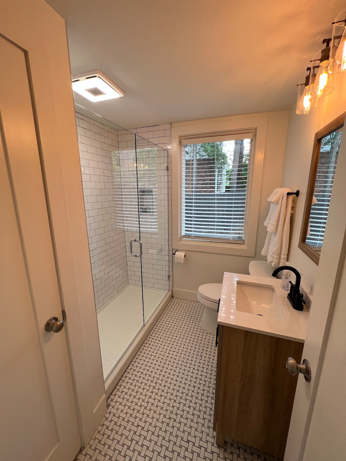 The bathroom features a glass shower enclosed with white subway tile, complemented by a modern sink with a wooden cabinet. A large window with white blinds allows natural light to enter, and a wall-mounted mirror is positioned above the sink.