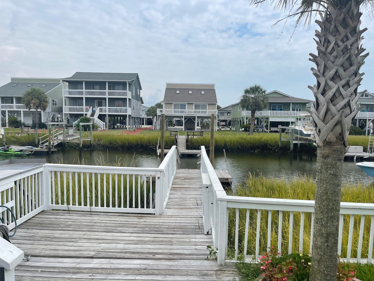 The image showcases a wooden dock extending over a calm waterway, with palmetto trees framing the view. Nearby, several houses are visible, nestled among tall grass along the banks of the canal. A subtle overcast sky contributes to the serene atmosphere.