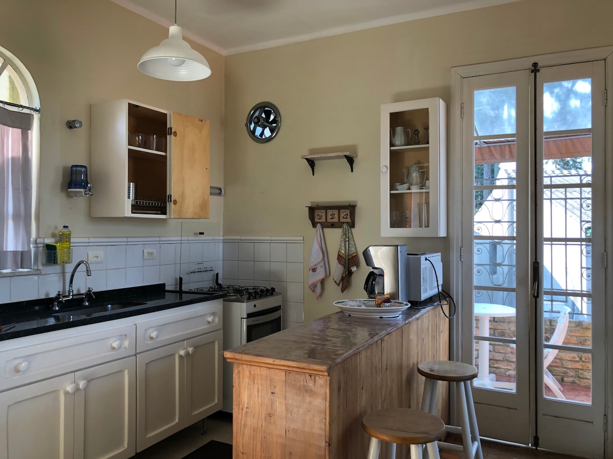 The kitchen area features a wooden island with two stools, along with white cabinetry and black countertops. Equipped with a gas stove and basic appliances, it opens to a balcony through glass doors, allowing natural light to fill the space.