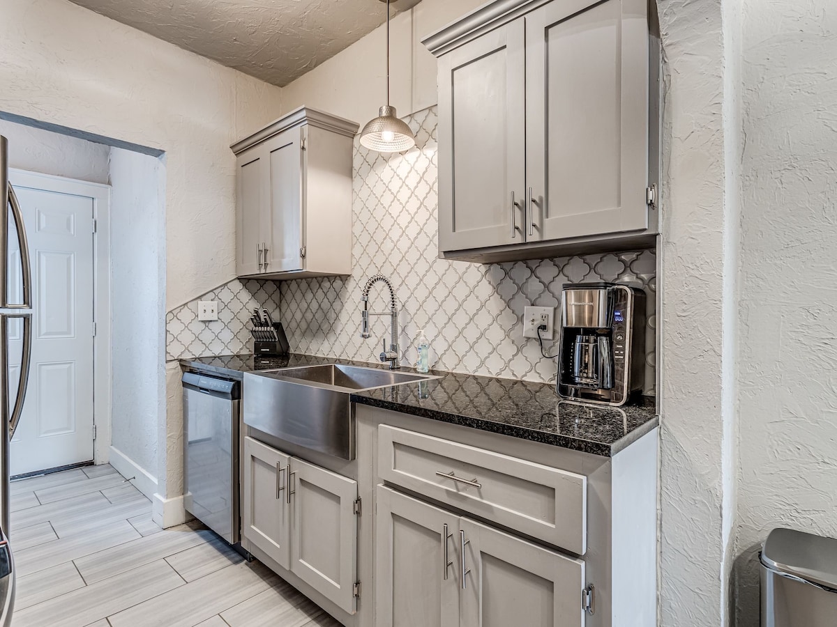 A modern kitchen features sleek gray cabinetry and a polished granite countertop. Stainless steel appliances, including a refrigerator and dishwasher, are present. A geometric tile backsplash provides texture, while ample lighting is offered by a pendant light hanging above the sink area.