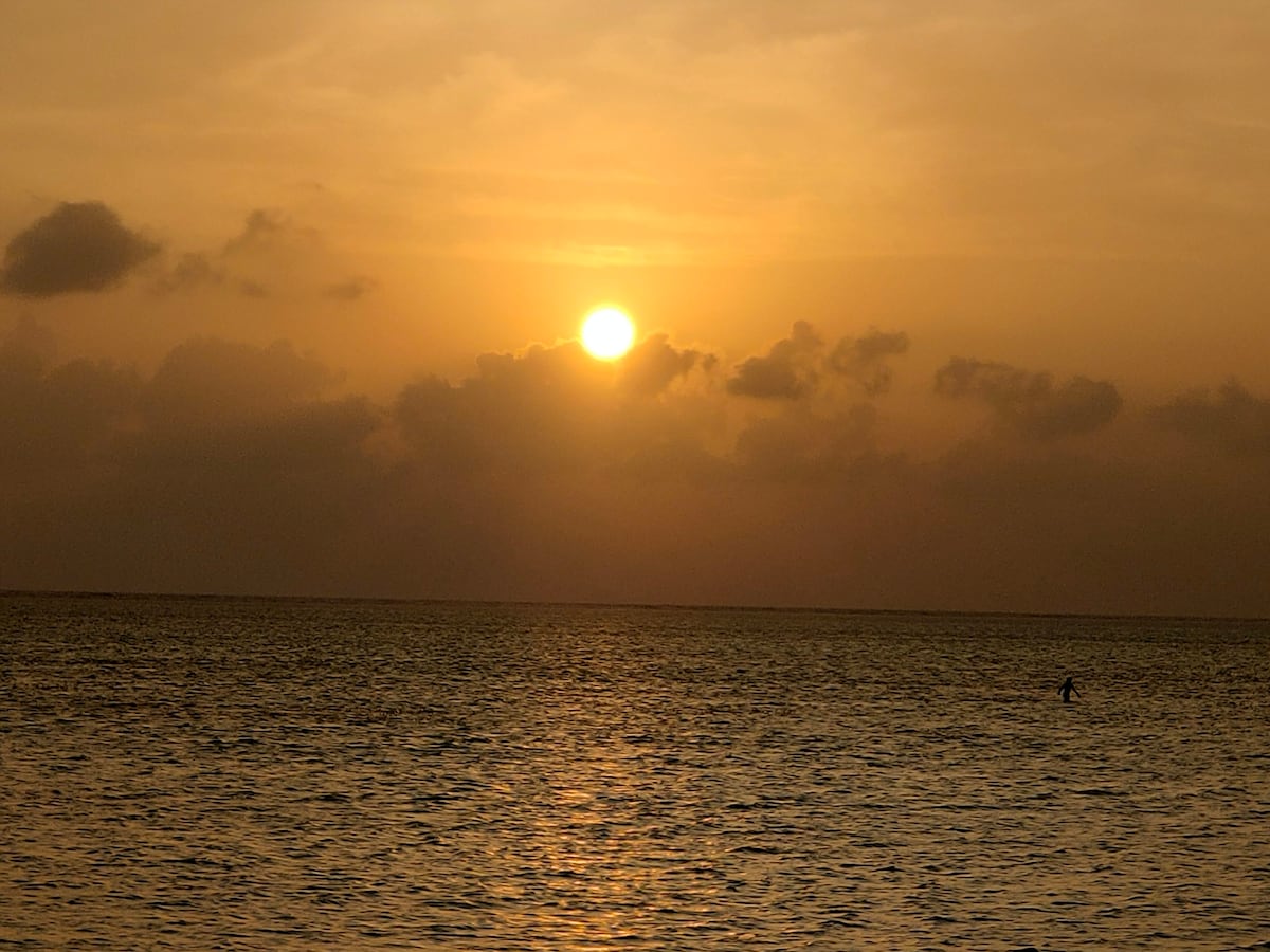 A serene sunset over the ocean is captured with warm hues of orange and soft clouds. The sun is partially submerged on the horizon, casting a golden reflection on the water's surface. A solitary figure is visible in the distance, standing in the water.