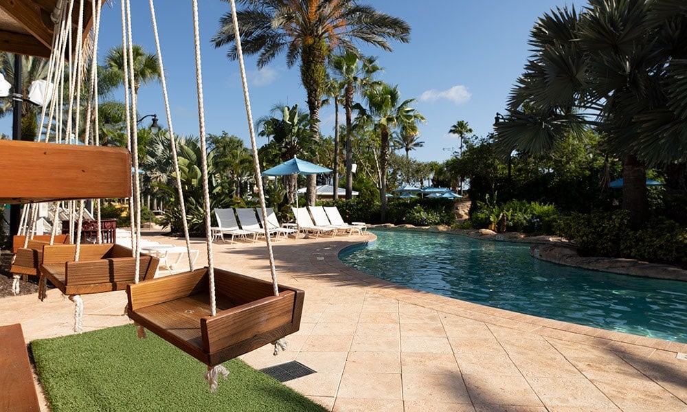 A serene pool area is surrounded by lush palm trees and providing a tranquil ambiance. Wooden swings are positioned near the pool, complementing the lounge chairs arranged under blue umbrellas. The sunlit water reflects the clear blue sky, enhancing the inviting atmosphere.