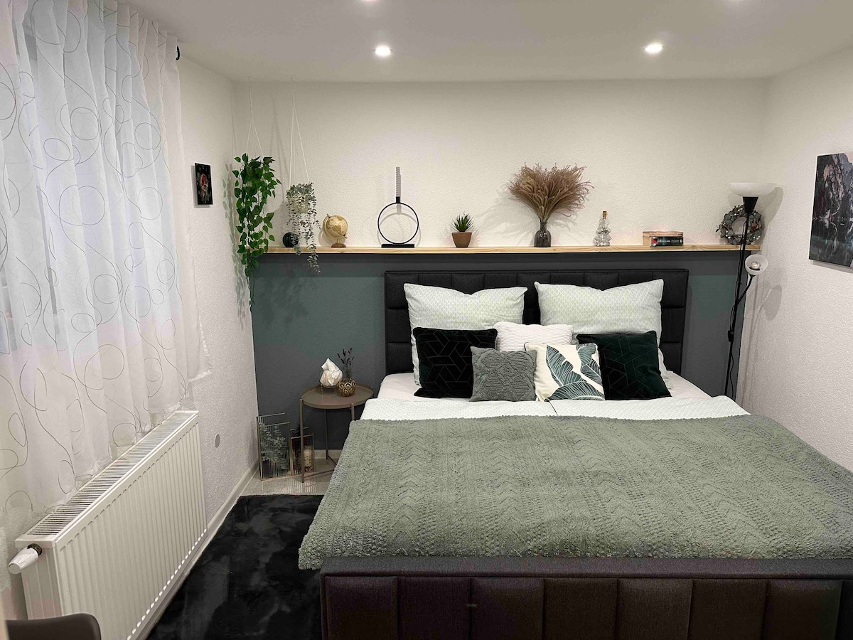 A cozy sleeping area is presented with a spacious bed adorned with a soft green throw and multiple decorative pillows. Natural light filters through sheer white curtains, complementing the calming decor, which includes small plants and tasteful accents on a nearby shelf.