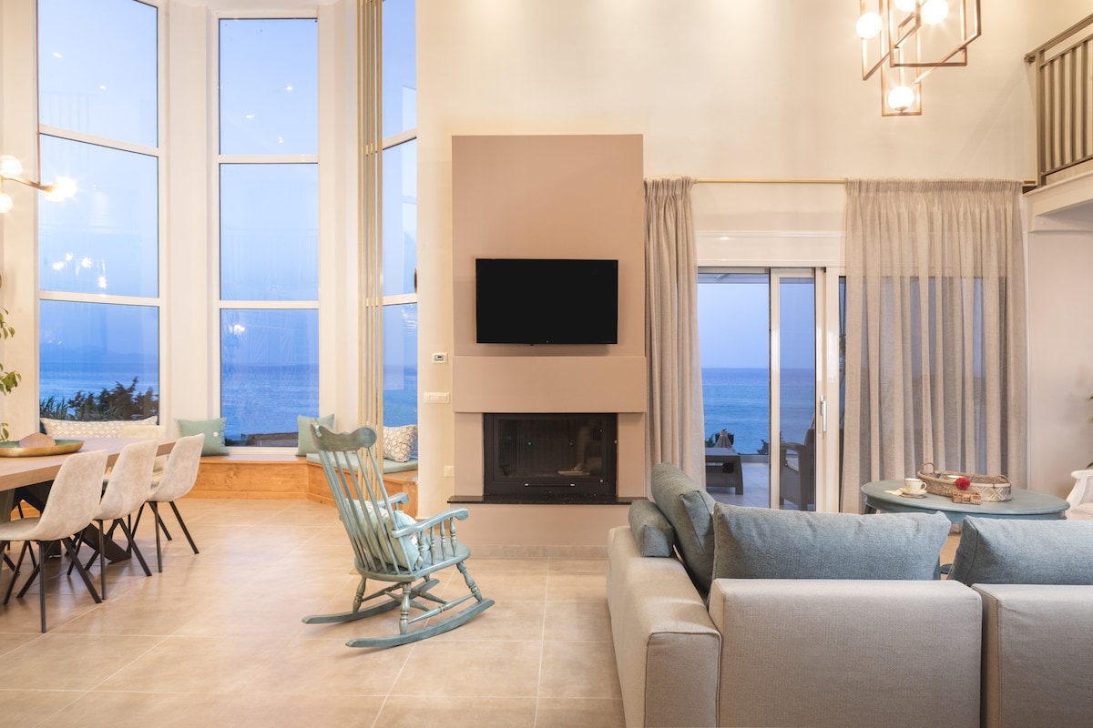 An airy living space features large windows offering unobstructed sea views. A light-colored sofa is arranged near a modern fireplace, while a rocking chair adds a cozy touch. A dining area with a spacious table is visible in the background, illuminated by soft lighting.