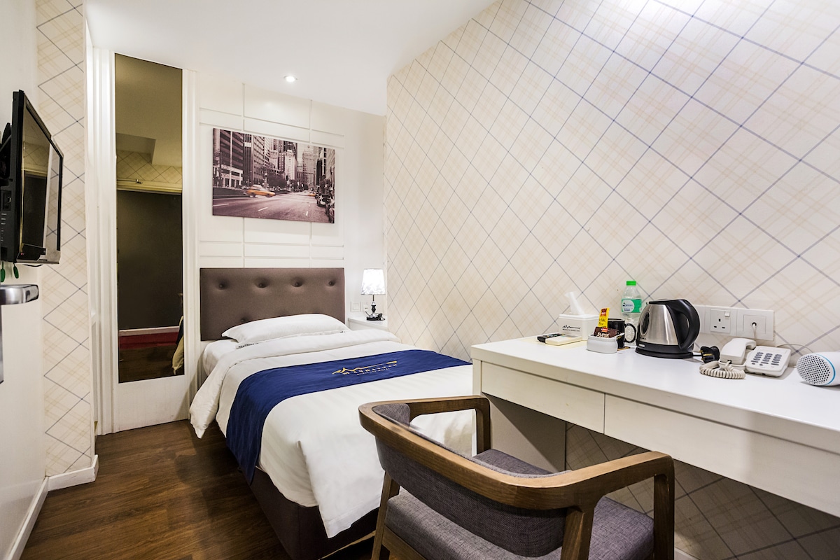 A compact room features a cozy bed adorned with a blue runner. A desk and chair are positioned next to a flat-screen television mounted on the wall. A kettle and various refreshments are displayed on the desk, while a large mirror reflects the light tones of the walls.