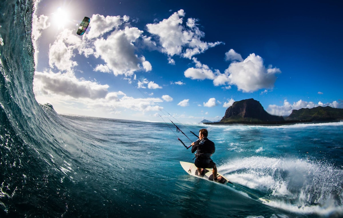 A kitesurfer is captured mid-flight above the waves, with a dramatic wave cresting nearby. The backdrop features a clear blue sky dotted with clouds and a lush mountain silhouette in the distance, illustrating the vibrant coastal environment.