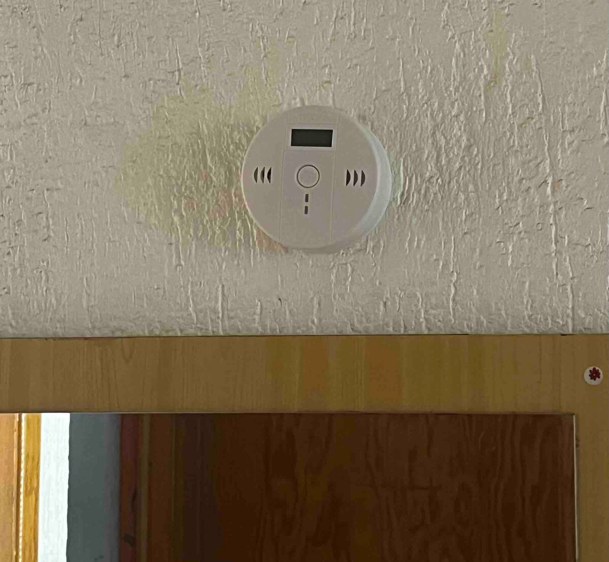 A smoke detector is mounted on a textured white ceiling, featuring a small digital display and several control buttons. The image also includes a wooden doorframe below, providing context to the room's design.