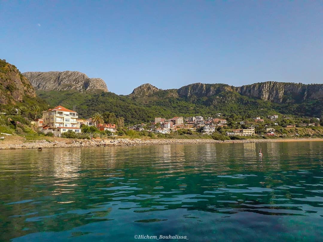 A serene coastal landscape is visible, featuring a clear blue water surface reflecting surrounding greenery. The shoreline is lined with a mix of buildings and vegetation, with a backdrop of rugged mountains under a clear sky.