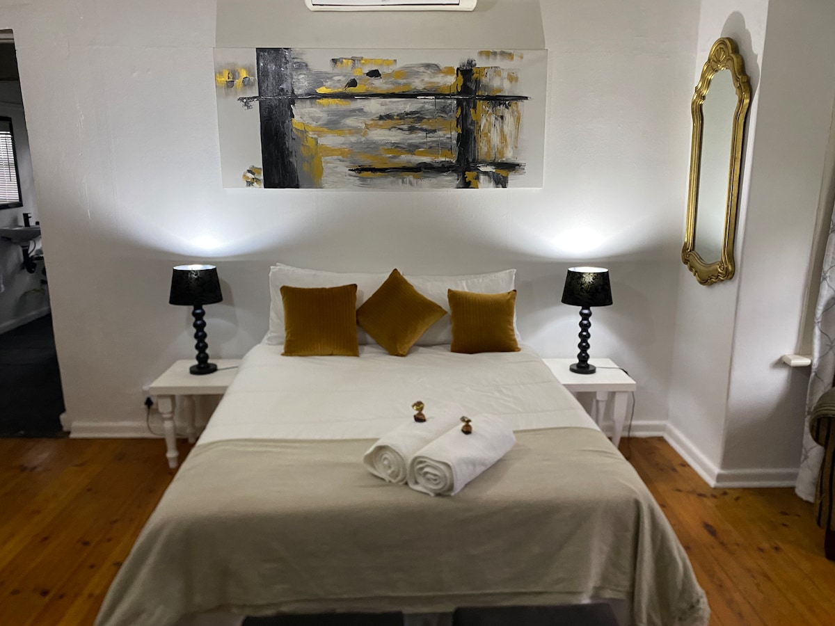A spacious bed is centered in the room, adorned with neutral linens and decorative pillows. Soft lighting from two bedside lamps creates a calm ambiance. An abstract painting in shades of black and gold is displayed above the bed, while a vintage mirror adds depth to the space.