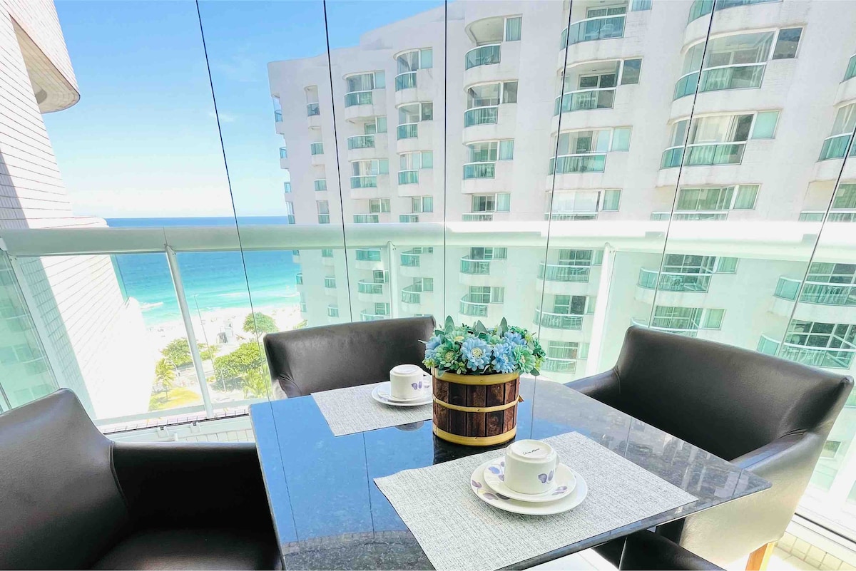 A glass-enclosed balcony features a small table set for two, adorned with a bouquet of blue flowers. Views of the ocean and nearby buildings are visible through the clear glass walls, creating an inviting space for relaxation.