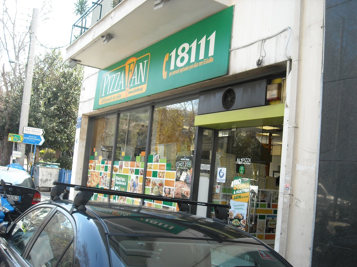 The image displays the exterior of a pizza restaurant, featuring a large storefront with glass windows showcasing colorful menu advertisements. A sign prominently displays the restaurant name, and a car is parked in front. Surrounding greenery and street signage are visible.