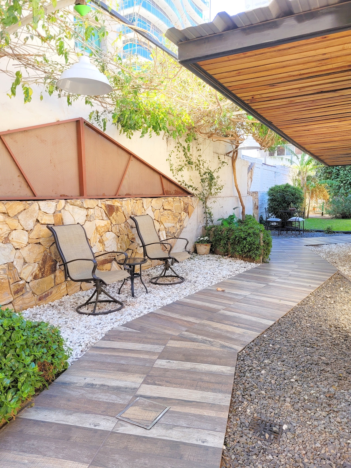A serene garden area features two chairs positioned on a pathway made of wooden planks. A decorative stone wall complements the lush greenery, with potted plants enhancing the natural setting. Sunlight filters through the surroundings, creating a quiet and welcoming outdoor space.