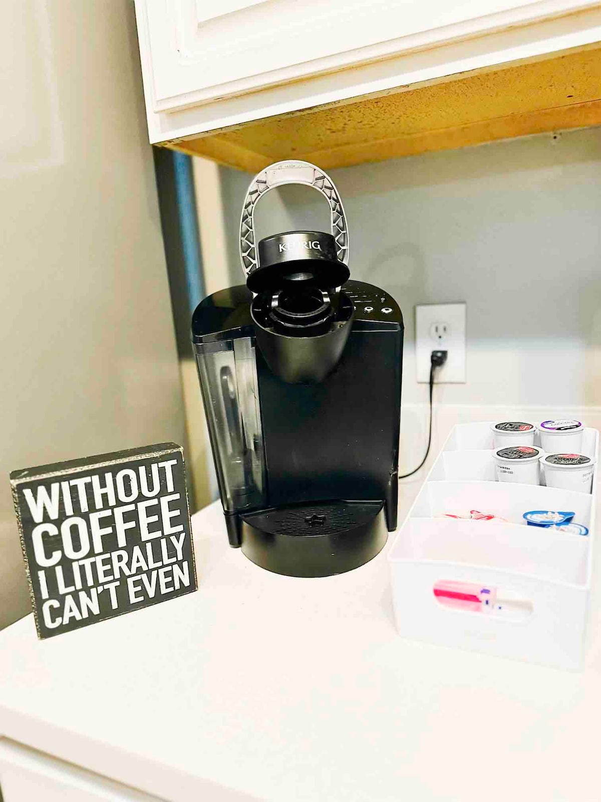 A coffee maker is positioned on a kitchen counter beside a small sign with a humorous text. A clear container holds coffee pods in various colors, while the coffee maker features an accessible design for ease of use.