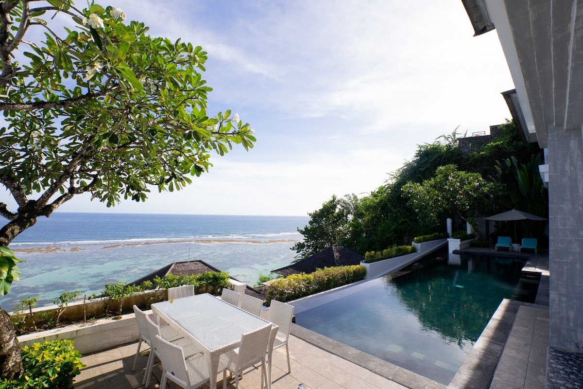 An outdoor dining area with a spacious table is accompanied by several white chairs. The infinity pool reflects the clear blue sky and ocean views, framed by lush greenery and a gentle slope. The tranquil setting invites relaxation, overlooking the serene beachfront.