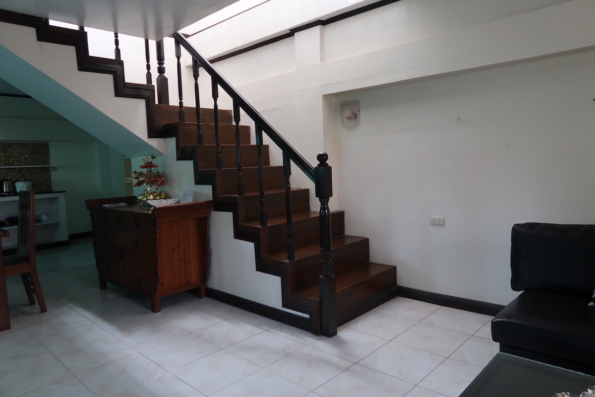 A wooden staircase leads to the second floor, enhanced by decorative bannisters. A small wooden cabinet sits against the wall near the stairs. The living area features a dark sofa, and neutral tiles cover the floor, contributing to the spacious feel of the ground level.