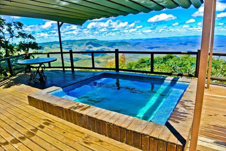 A wooden deck features a private jacuzzi, bordered by elegant tile work. The structure is partially shaded by an overhang, providing a retreat to enjoy the expansive mountain views. The surrounding area is lush, with trees and sweeping vistas of the landscape.