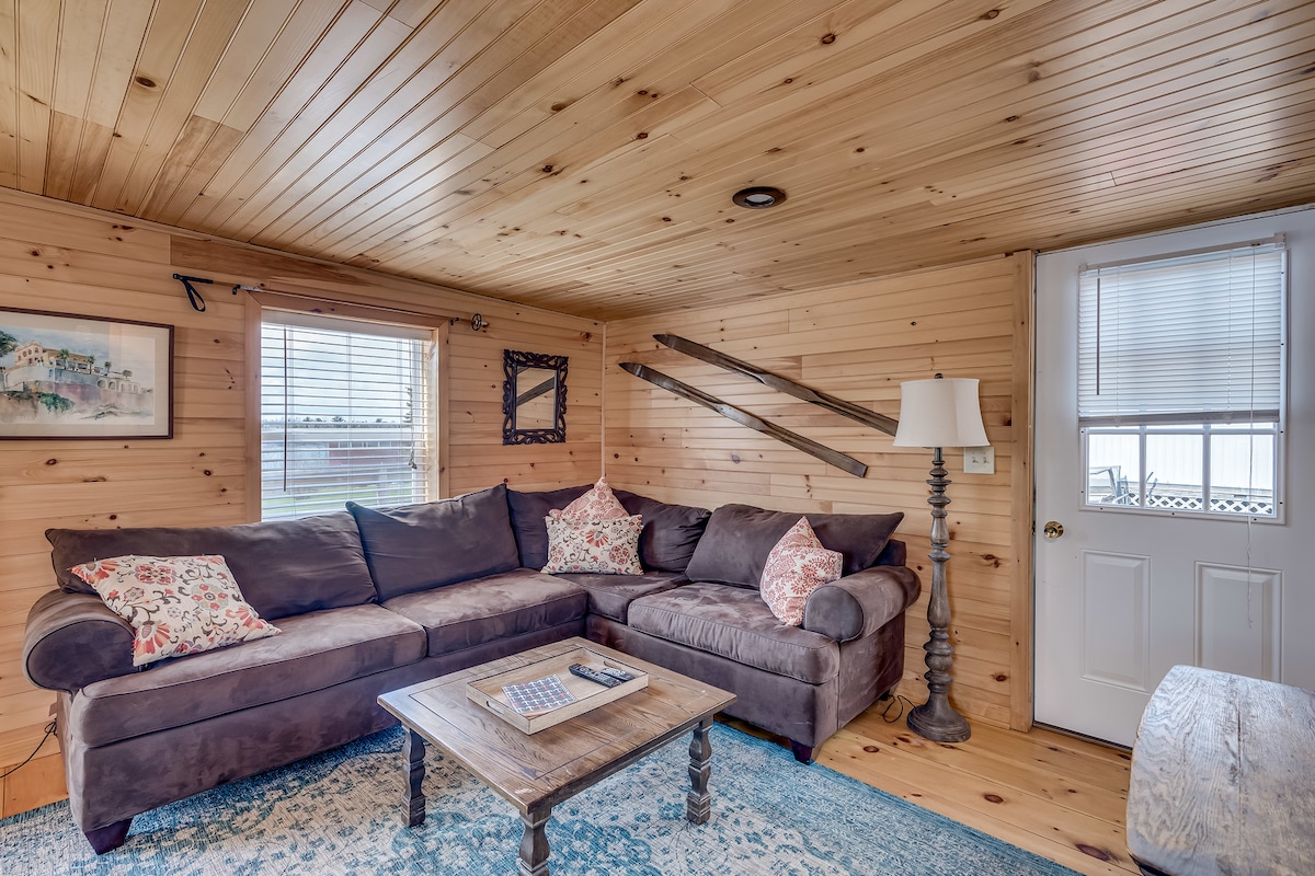 A cozy living area is surrounded by warm wooden walls, featuring a large, cushioned sectional sofa adorned with decorative pillows. A small coffee table sits in front, topped with a book. Natural light filters through the windows, illuminating the inviting space.