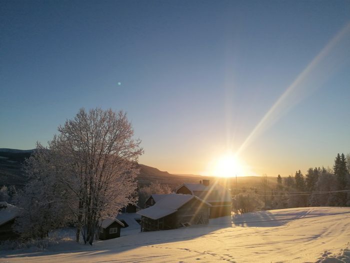 A serene winter landscape is visible, showcasing a sun setting over snow-covered hills. Trees with frosted branches stand nearby, adding contrast to the scene. Several cabins are nestled within the snow, creating a cozy atmosphere amidst the tranquil environment.