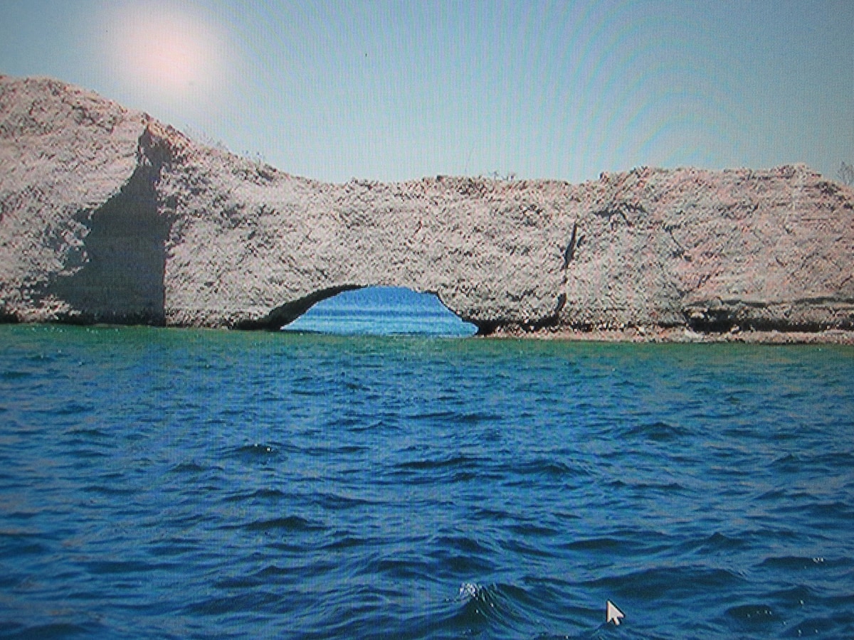 A natural rock formation rises from the water, featuring a prominent arch that creates a passage through the structure. The surrounding water reflects shades of blue and green, contrasting with the rocky surface. The distant horizon hints at additional land beyond the arch.