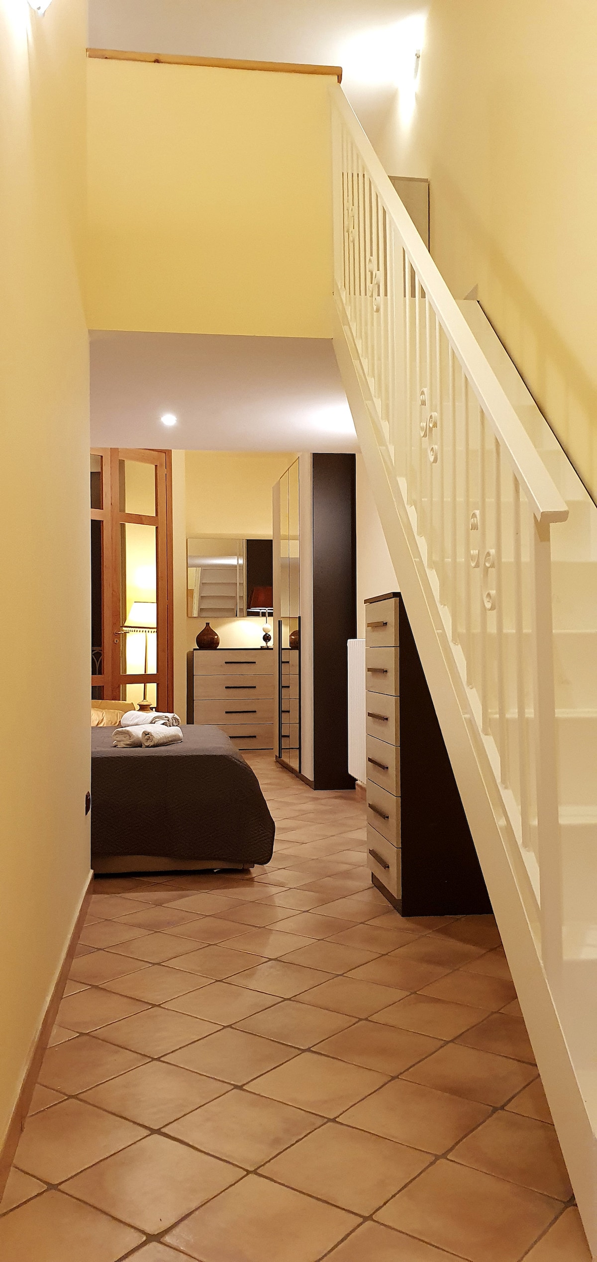 A well-lit corridor leads towards a staircase. To the left, a cozy sleeping area features a neatly made bed with a grey coverlet, accompanied by a dresser and decorative accents. Warm yellow walls enhance the inviting atmosphere, while tiles cover the floor for easy navigation.