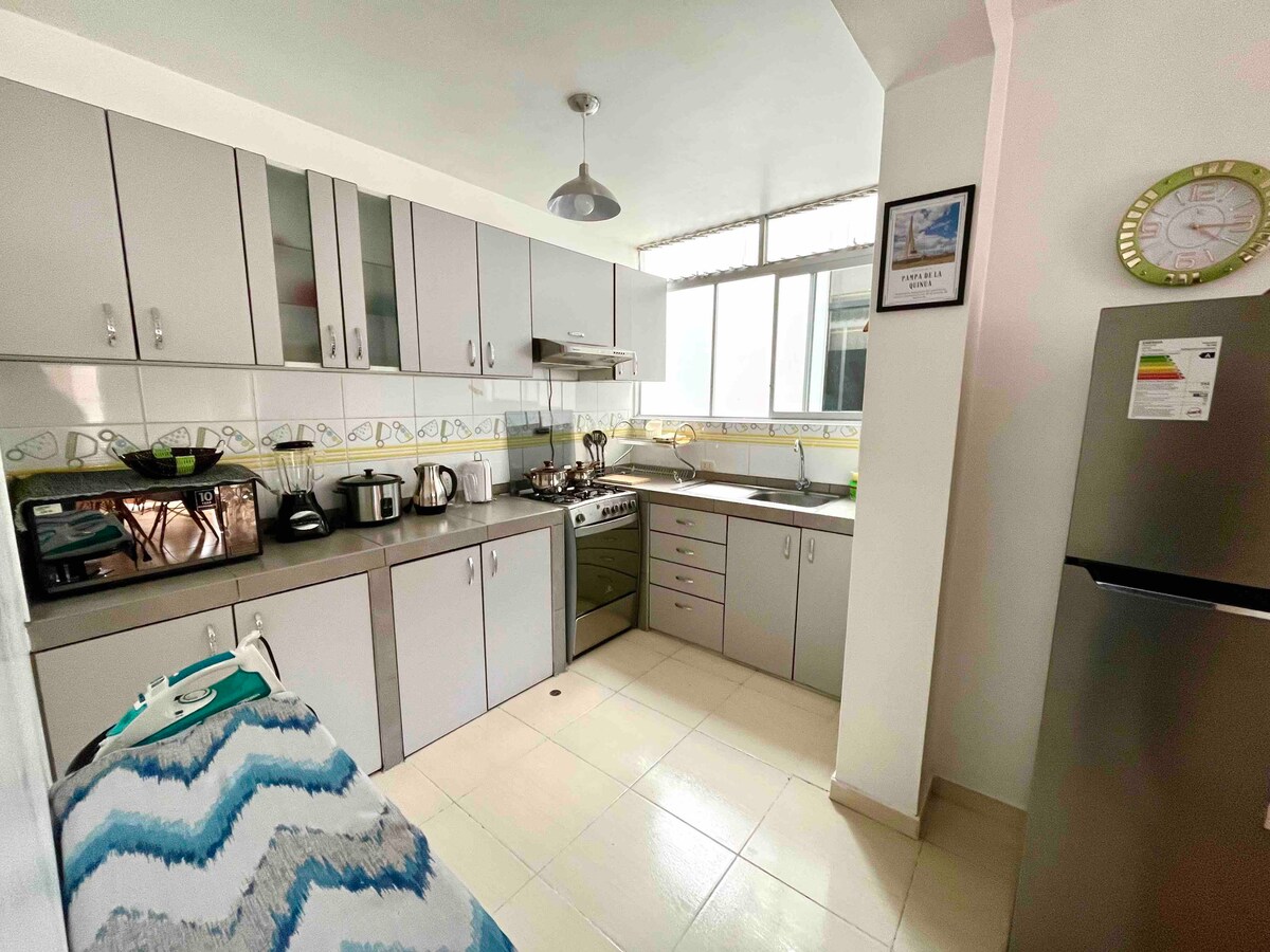 A modern kitchen is equipped with silver cabinetry, a gas stove, and essential appliances, including a microwave and kettle. Natural light fills the space through a large window. A refrigerator stands against one wall, while a patterned towel is neatly placed on the countertop.