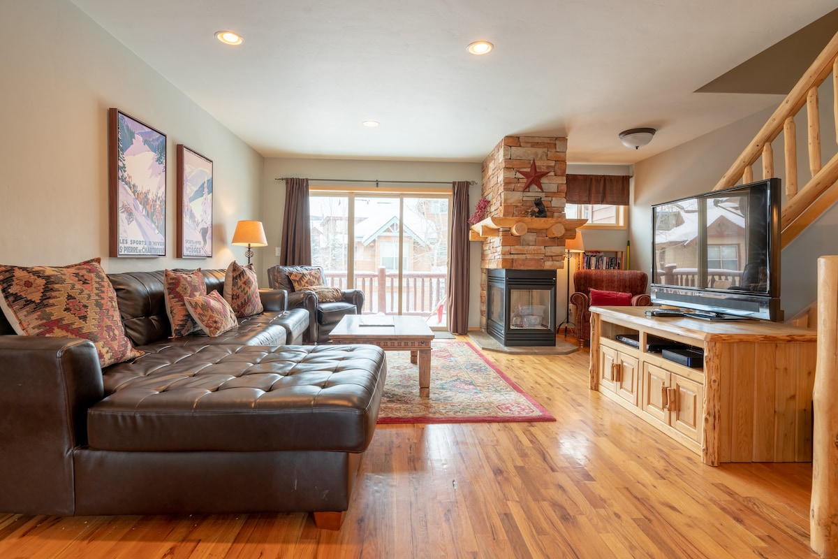 The living area features a spacious leather sectional sofa arranged around a coffee table, creating an inviting space. A stone fireplace serves as a focal point, while large windows allow natural light to fill the room. A television is positioned within a wooden console.