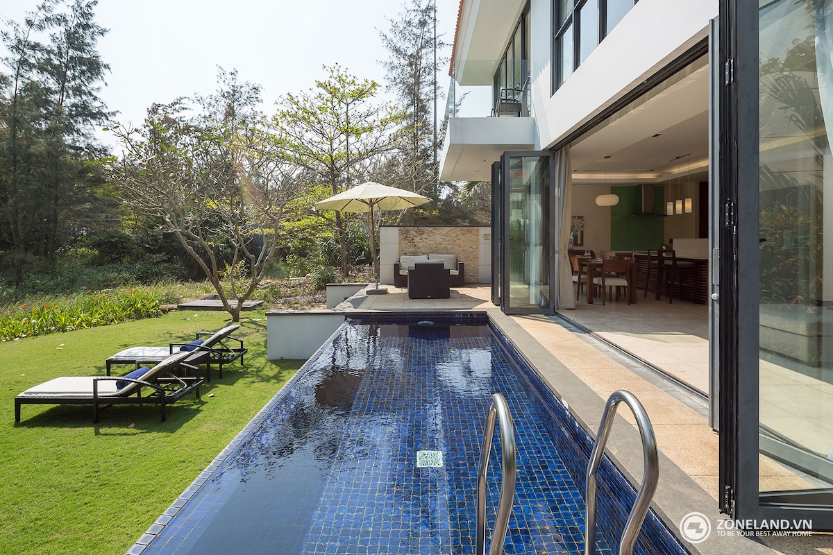 A private plunge pool is framed by a manicured lawn and tropical greenery. Expansive glass doors create an open transition between indoor and outdoor spaces. A sun umbrella provides shade over a seating area, enhancing a tranquil setting suitable for relaxation.
