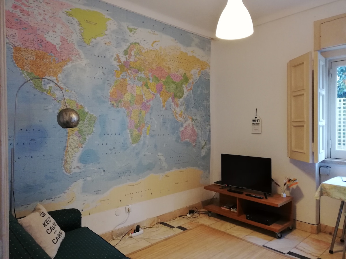 A spacious living area features a large world map mural on one wall, providing a vibrant backdrop. A green sofa is positioned nearby, complemented by a modern floor lamp. A wooden TV stand supports a flat-screen television, while natural light enters through a window with beige shutters.