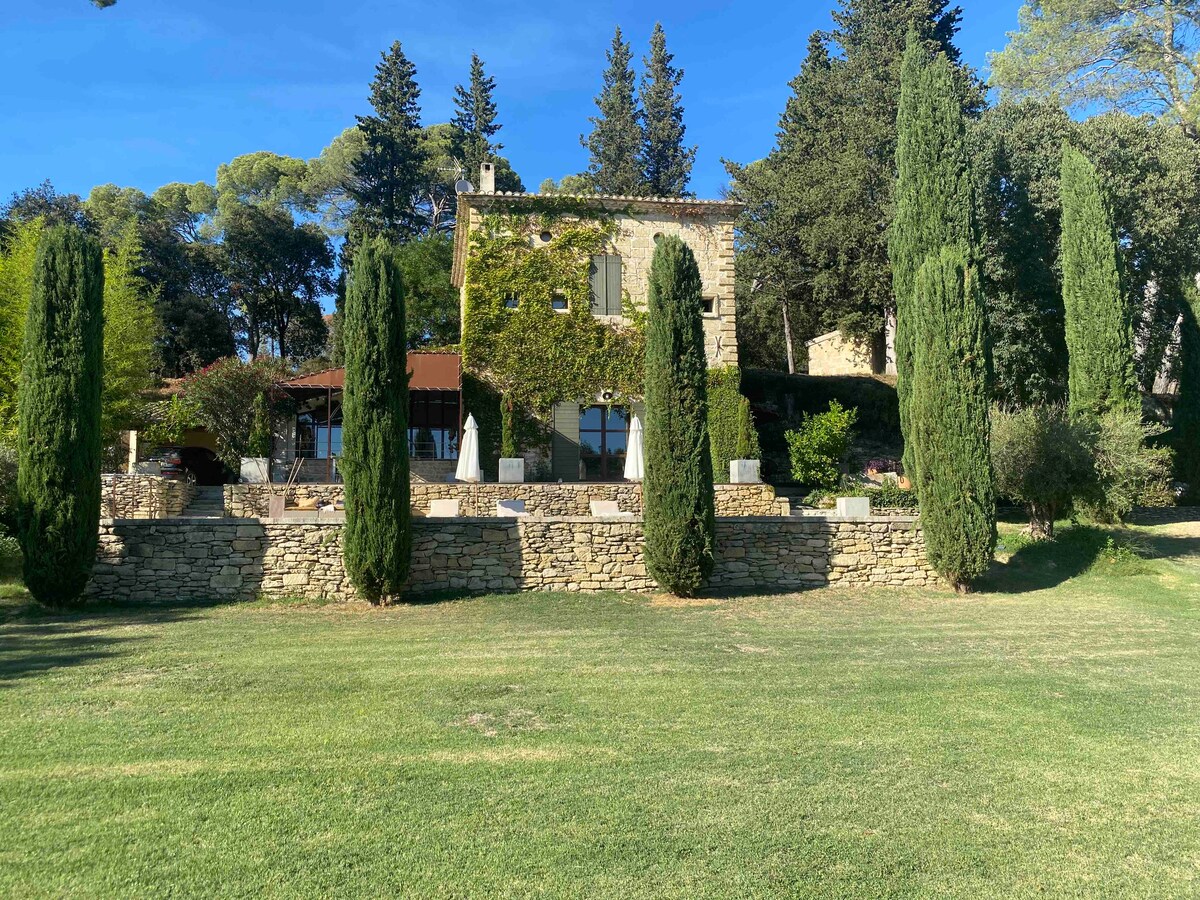 A charming stone building is set amidst verdant greenery, framed by tall, slender cypress trees. The landscaped yard features neatly trimmed grass and stone retaining walls that enhance the natural beauty of the setting.